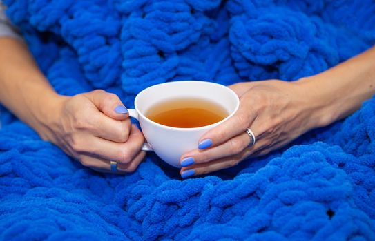 A girl wrapped in a large blue merino wool hand knitted blanket, a warm and very soft blanket, a girl holding a white cup of hot tea, place for text