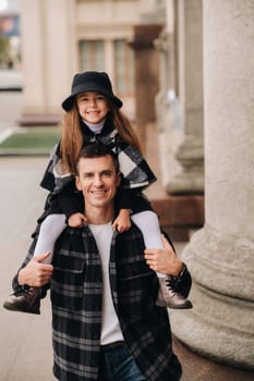 A cheerful Daughter is sitting on dad's shoulders and walking around the autumn city.