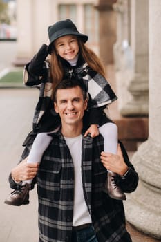 A cheerful Daughter is sitting on dad's shoulders and walking around the autumn city.