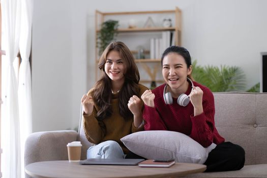 Two Young womanWatching TV Shaking Fists In Joy Celebrating Victory Of Favorite Sport Team Sitting On Couch In Living Room At Home. Weekend Leisure, Television Show And Entertainment Concept.