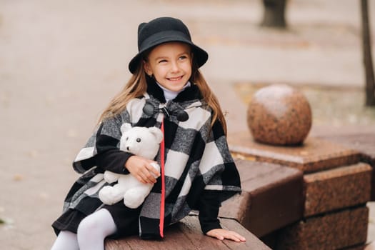 A little stylish girl in autumn clothes is sitting on a bench in the autumn city.