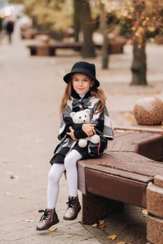 A little stylish girl in autumn clothes is sitting on a bench in the autumn city.