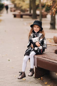 A little stylish girl in autumn clothes is sitting on a bench in the autumn city.
