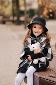 A little stylish girl in autumn clothes is sitting on a bench in the autumn city.