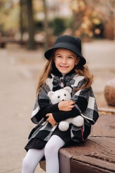 A little stylish girl in autumn clothes is sitting on a bench in the autumn city.