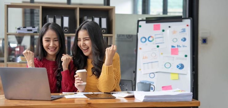 Two young Asian businesswomen show joyful expression of success at work smiling happily with a laptop computer in a modern office..