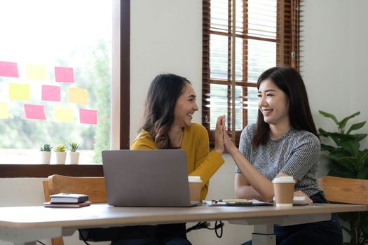 Two young Asian businesswomen show joyful expression of success at work smiling happily with a laptop computer in a modern office..