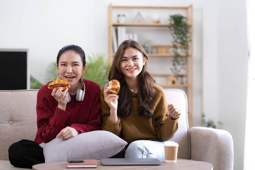 Two Young womanWatching TV Shaking Fists In Joy Celebrating Victory Of Favorite Sport Team Sitting On Couch In Living Room At Home. Weekend Leisure, Television Show And Entertainment Concept.