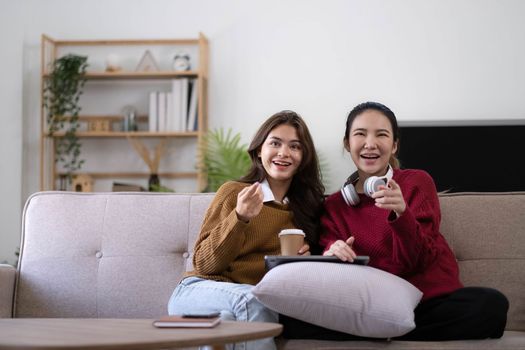 Two Young womanWatching TV Shaking Fists In Joy Celebrating Victory Of Favorite Sport Team Sitting On Couch In Living Room At Home. Weekend Leisure, Television Show And Entertainment Concept.