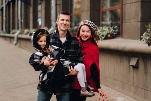 A stylish family of three people are walking in the city in the fall and dad is holding his daughter in his arms.