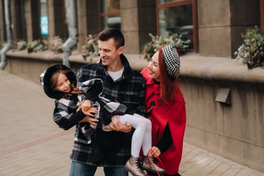 A stylish family of three people are walking in the city in the fall and dad is holding his daughter in his arms.