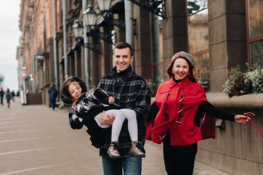 A stylish family of three people are walking in the city in the fall and dad is holding his daughter in his arms.