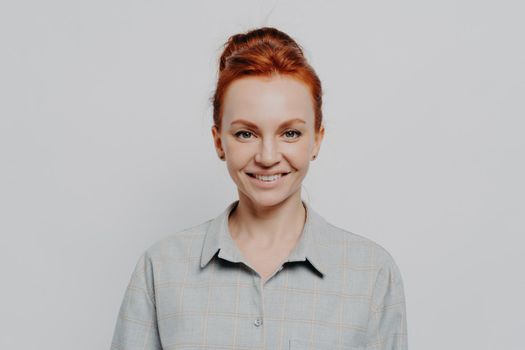 Headshot portrait of happy young caucasian ginger woman being satisfied and feeling happy, looking at camera cheerful while standing isolated on grey background. Positive emotions concept