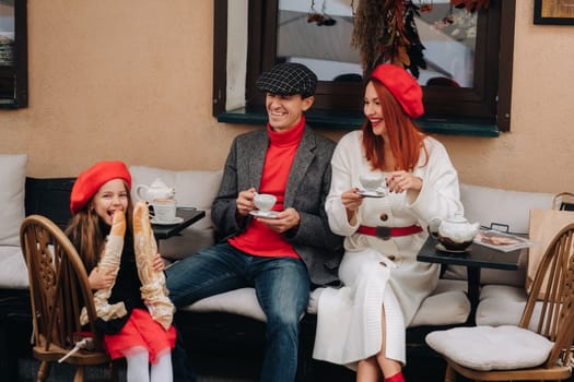 A stylish family of three is sitting at a table outside in a cafe and drinking coffee. Dad, mom and daughter in the autumn city.