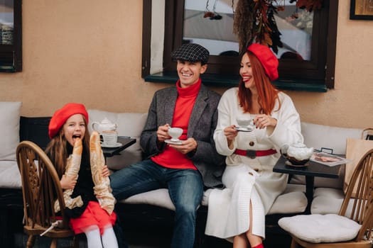 A stylish family of three is sitting at a table outside in a cafe and drinking coffee. Dad, mom and daughter in the autumn city.