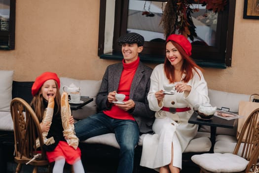 A stylish family of three is sitting at a table outside in a cafe and drinking coffee. Dad, mom and daughter in the autumn city.