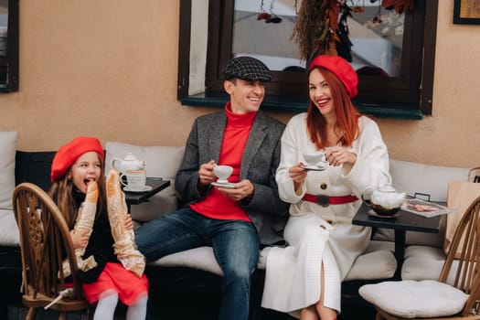 A stylish family of three is sitting at a table outside in a cafe and drinking coffee. Dad, mom and daughter in the autumn city.