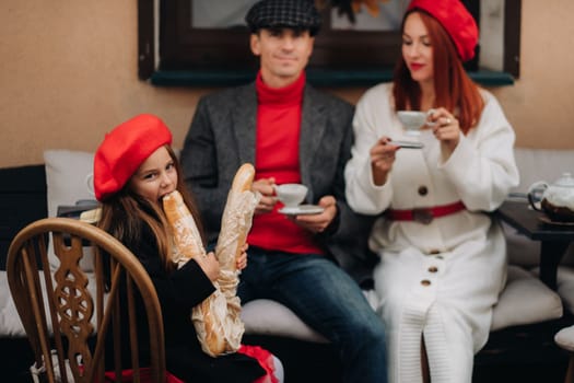 A stylish family of three is sitting at a table outside in a cafe and drinking coffee. Dad, mom and daughter in the autumn city.