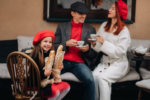 A stylish family of three is sitting at a table outside in a cafe and drinking coffee. Dad, mom and daughter in the autumn city.