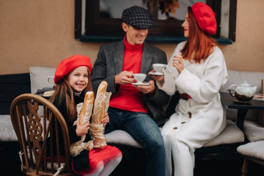 A stylish family of three is sitting at a table outside in a cafe and drinking coffee. Dad, mom and daughter in the autumn city.