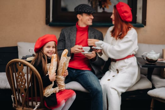 A stylish family of three is sitting at a table outside in a cafe and drinking coffee. Dad, mom and daughter in the autumn city.