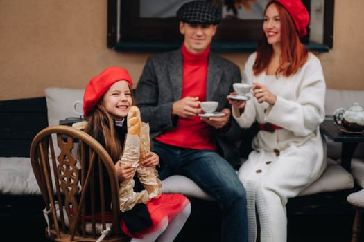 A stylish family of three is sitting at a table outside in a cafe and drinking coffee. Dad, mom and daughter in the autumn city.