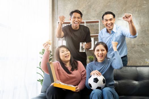 Friends watch sports on TV, cheer and celebrate. Happy diverse asian friend supporters fans sit on couch with popcorn and drinks.