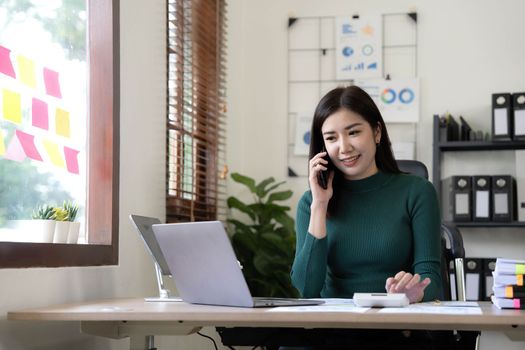 Smiling beautiful Asian businesswoman analyzing chart and graph showing changes on the market and holding smartphone at office..