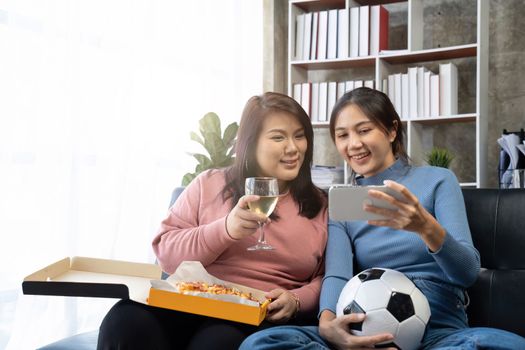 Friends watch sports on mobile phone, cheer and celebrate. Happy diverse asian friend supporters fans sit on couch with popcorn and drinks.