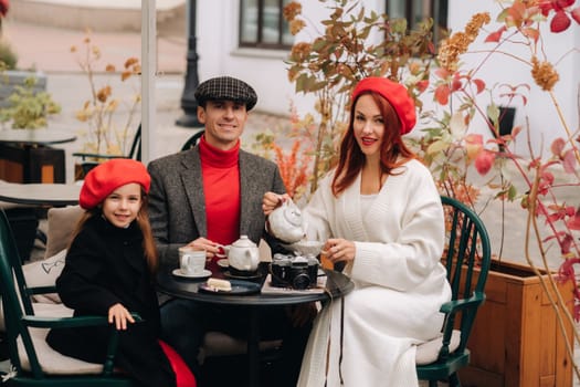 A stylish family gathered together in a cafe on the street. Mom, dad, little daughter drink tea, eat cakes. They are happy together. The concept of a happy family dinner.