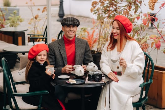 A stylish family gathered together in a cafe on the street. Mom, dad, little daughter drink tea, eat cakes. They are happy together. The concept of a happy family dinner.