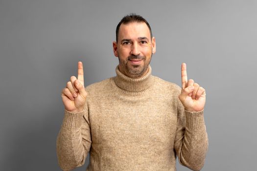 Portrait of a bearded latino man in turtleneck sweater happy pointing fingers at copyspace isolated over gray background