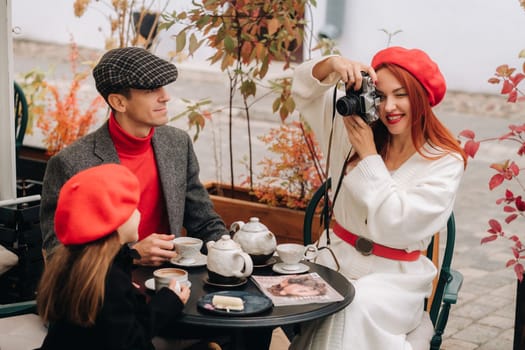 A stylish family of three is sitting at a table outside in a cafe and drinking coffee. Dad, mom and daughter in the autumn city.