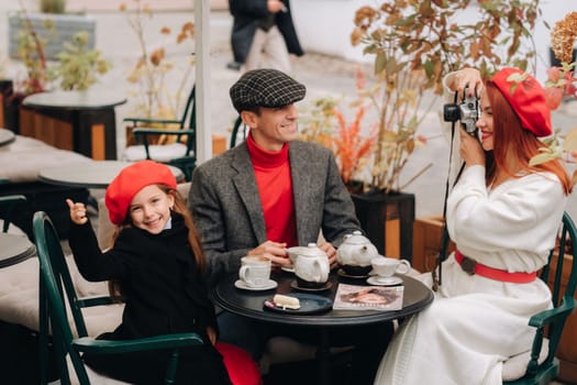 A stylish family of three is sitting at a table outside in a cafe and drinking coffee. Dad, mom and daughter in the autumn city.