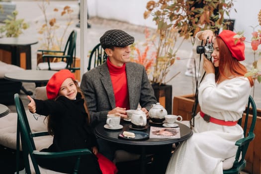 A stylish family of three is sitting at a table outside in a cafe and drinking coffee. Dad, mom and daughter in the autumn city.