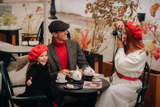 A stylish family of three is sitting at a table outside in a cafe and drinking coffee. Dad, mom and daughter in the autumn city.