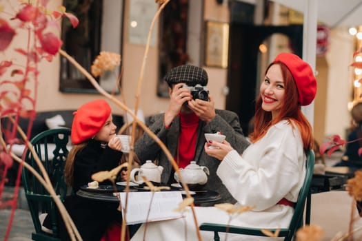 A stylish family of three is sitting at a table outside in a cafe and drinking coffee. Dad takes pictures of mom in the autumn city.