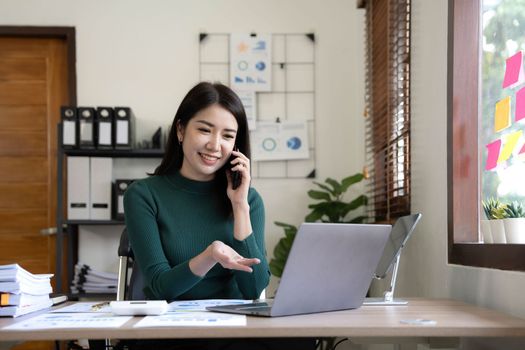 Smiling beautiful Asian businesswoman analyzing chart and graph showing changes on the market and holding smartphone at office..