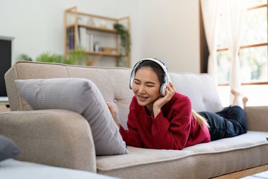 Carefree asian woman listening to music, using headphones, laying on couch in living room, copy space. Happy young lady with closed eyes enjoying music, home interior.
