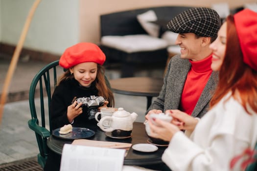 A stylish family of three is sitting at a table outside in a cafe and drinking coffee. Dad, mom and daughter in the autumn city.