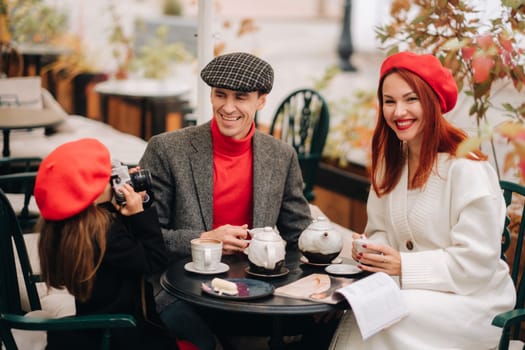 A stylish family of three is sitting at a table outside in a cafe and drinking coffee. Dad, mom and daughter in the autumn city.