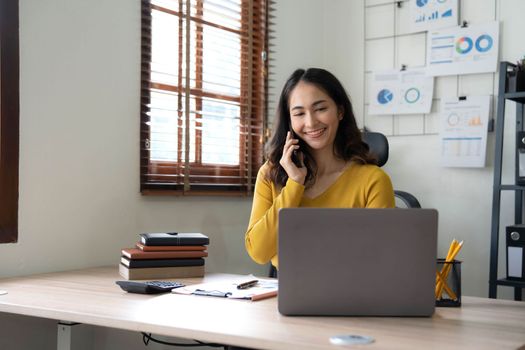 Smiling beautiful Asian businesswoman analyzing chart and graph showing changes on the market and holding smartphone at office..