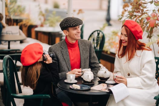 A stylish family of three is sitting at a table outside in a cafe and drinking coffee. Dad, mom and daughter in the autumn city.