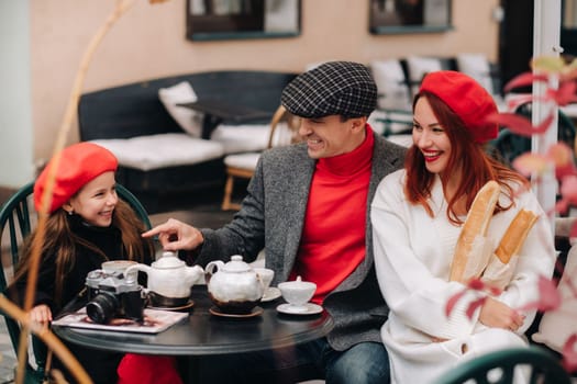 A stylish family gathered together in a cafe on the street. Mom, dad, little daughter drink tea, eat cakes. They are happy together. The concept of a happy family dinner.