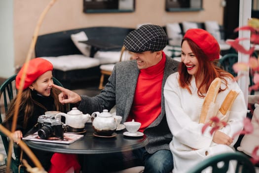 A stylish family gathered together in a cafe on the street. Mom, dad, little daughter drink tea, eat cakes. They are happy together. The concept of a happy family dinner.