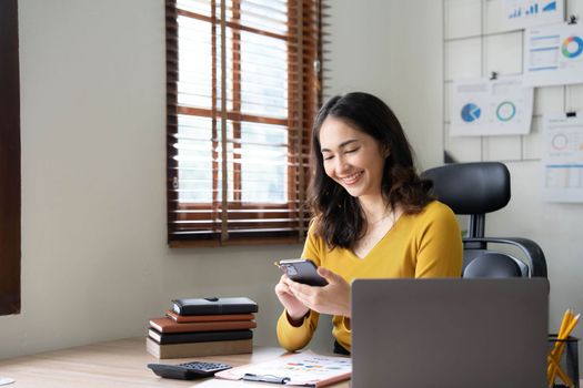 Smiling beautiful Asian businesswoman analyzing chart and graph showing changes on the market and holding smartphone at office..