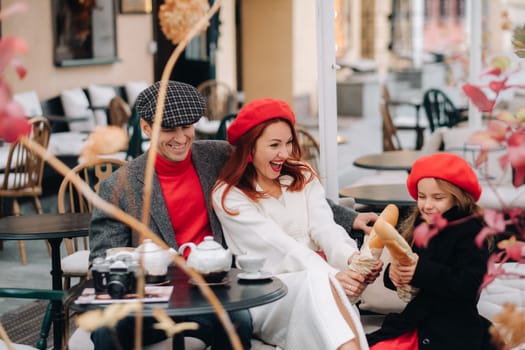 A stylish family of three is sitting at a table outside in a cafe and drinking coffee. Dad, mom and daughter in the autumn city.