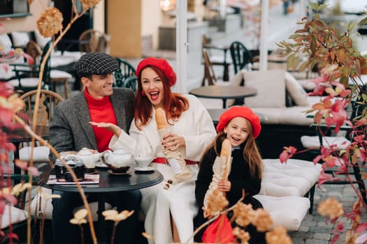 A stylish family of three is sitting at a table outside in a cafe and drinking coffee. Dad, mom and daughter in the autumn city.