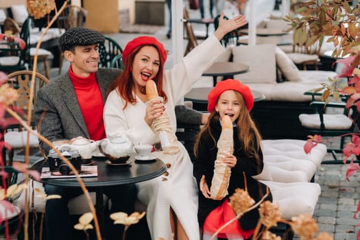 A stylish family of three is sitting at a table outside in a cafe and drinking coffee. Dad, mom and daughter in the autumn city.