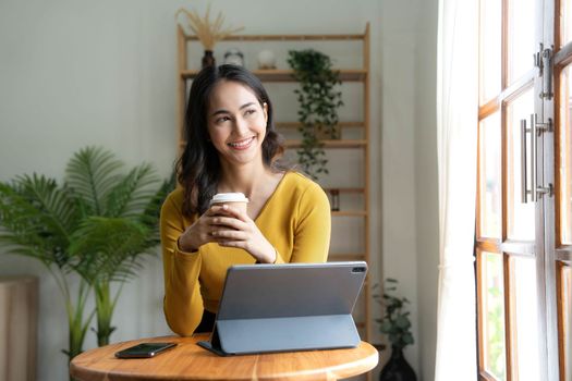 Beautiful Asian woman is using laptop, drinking coffee and smiling while sitting by the window at home...